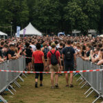 A crowded outdoor festival scene with numerous people standing behind metal barricades, with tents and trees in the background.