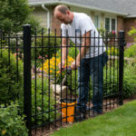 A man working in a garden, using a tool near a black metal fence surrounded by colorful flowers, with a house visible in the background.