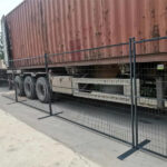 A close-up view of a wire mesh fence alongside a large shipping container truck, highlighting the fence's structure and the vehicle's wheels. The truck is parked on a concrete surface.