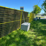 A yellow wire mesh fence secured to concrete blocks, surrounding a grassy area, with a clear blue sky and a sign visible in the background, indicating a commercial space.