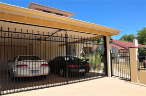 A black metal gate is shown in front of a carport where two cars are parked. The gate features decorative spikes and is positioned under an awning, with a clear blue sky in the background.