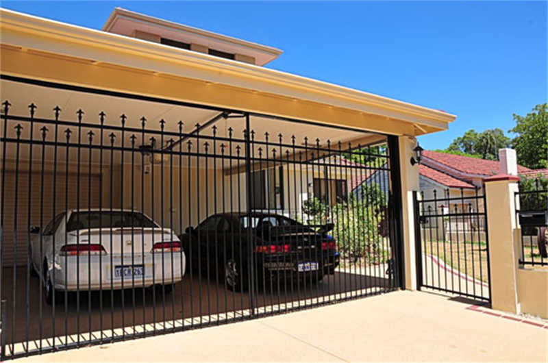 A black metal gate is shown in front of a carport where two cars are parked. The gate features decorative spikes and is positioned under an awning, with a clear blue sky in the background.