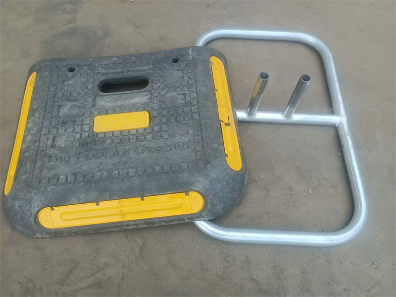 A close-up of a heavy-duty plastic base with yellow edges next to an aluminum frame, which is used to support fencing, lying on a sandy surface.