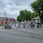 A street scene shows temporary fencing set up along the sidewalk, guiding pedestrians as people prepare for an event. Shops and trees line the street, with a cloudy sky above.