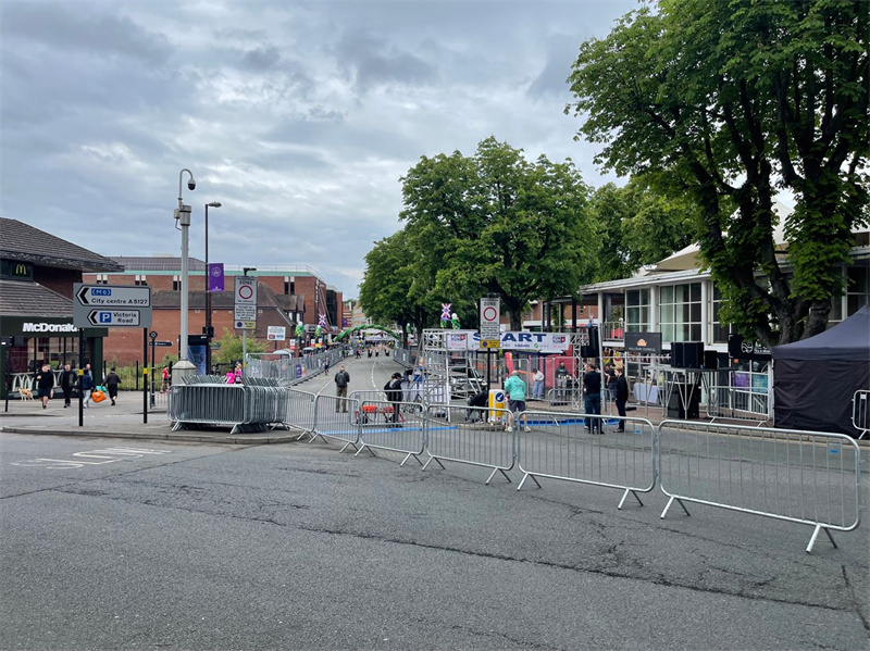 A street scene shows temporary fencing set up along the sidewalk, guiding pedestrians as people prepare for an event. Shops and trees line the street, with a cloudy sky above.