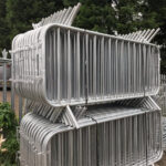 A close-up view of stacked metal crowd control barriers, showing multiple layers piled on top of each other. The barriers are shiny and made of metal, with some greenery visible in the background.
