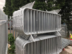 A close-up view of stacked metal crowd control barriers, showing multiple layers piled on top of each other. The barriers are shiny and made of metal, with some greenery visible in the background.