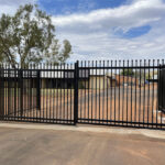 A large black metal gate with vertical bars and decorative tops, set in an open position, leading to a dirt driveway with trees and buildings in the background.