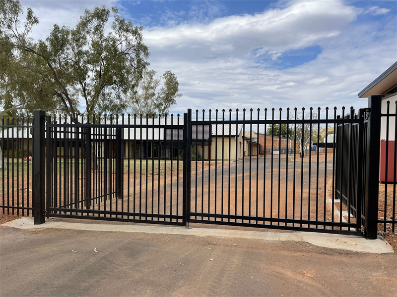 A large black metal gate with vertical bars and decorative tops, set in an open position, leading to a dirt driveway with trees and buildings in the background.