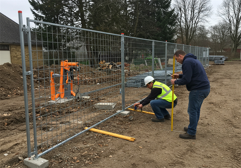 Two workers are adjusting a temporary fence on a construction site, with one measuring the fence alignment while the other secures it, surrounded by construction debris and materials.