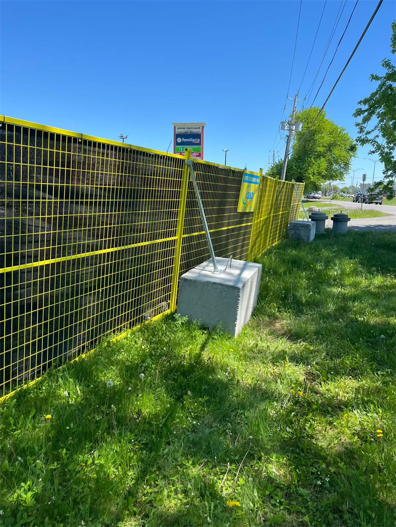 A yellow wire mesh fence secured to concrete blocks, surrounding a grassy area, with a clear blue sky and a sign visible in the background, indicating a commercial space.