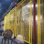 An indoor manufacturing space with several yellow construction fence panels hanging from a rack. Workers are visible in the background, and a fan is positioned nearby.