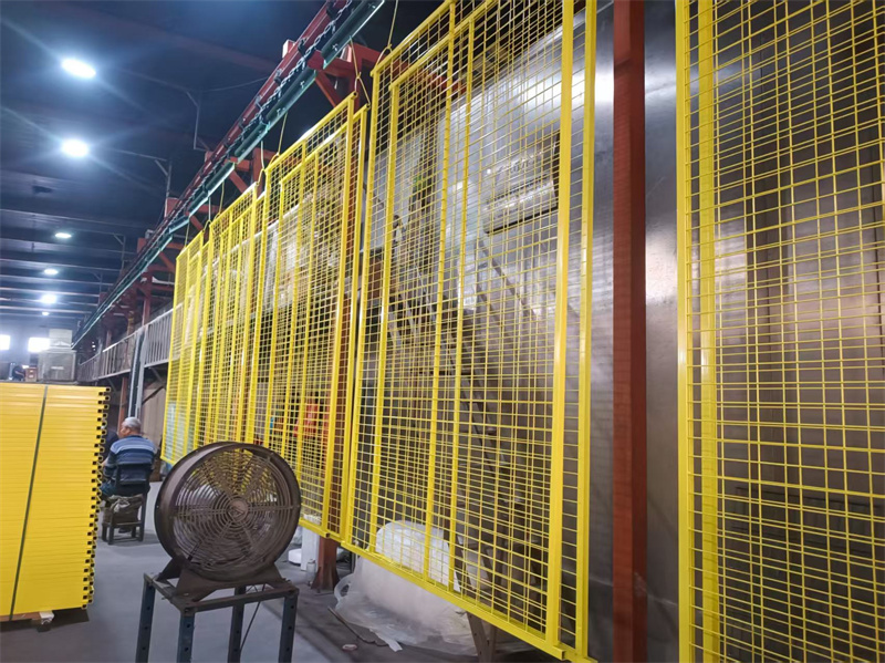 An indoor manufacturing space with several yellow construction fence panels hanging from a rack. Workers are visible in the background, and a fan is positioned nearby.