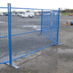 A blue construction fence with a gate, positioned on a gravel surface in an outdoor area, with parked vehicles visible in the background.