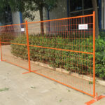 An orange construction fence set up outdoors, with a green hedge in the background. The fence has a label attached and is installed on a concrete surface.