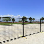 A section of black metal fencing secured to the ground, overlooking a vacant area under a clear blue sky.