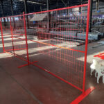 A red construction fence set up indoors, with bright lighting illuminating the space. A small white dog is visible in the foreground, and various construction materials are scattered around.