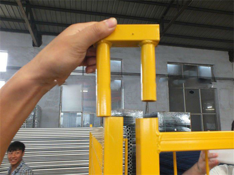 A close-up of a person holding a yellow fence connector, demonstrating how it fits into a metal fence panel, with stacks of fencing materials visible in the background.