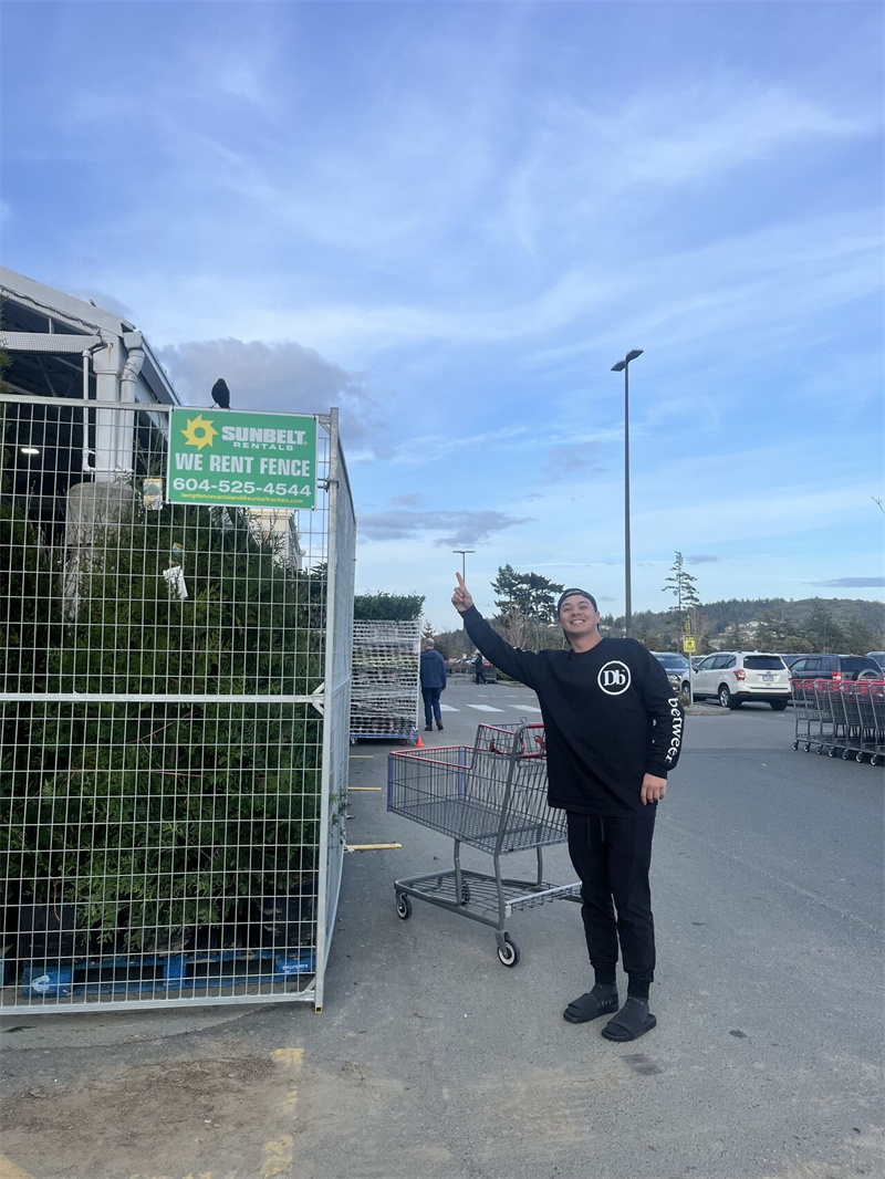 A person in a black sweatshirt standing next to a green fence with a sign that reads "We Rent Fence" from "Sunbelt Rentals," gesturing towards the sign while shopping carts are visible in the background.