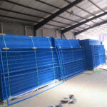 A blue construction fence surrounding a construction site, with equipment and materials visible inside the fenced area, indicating an active work zone.