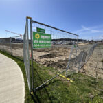 A construction site fenced off with a chain-link fence displaying a "Sunbelt Rentals We Rent Fence" sign. The area is cleared, with machinery and buildings visible in the background under a clear blue sky.