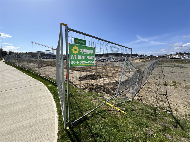 A construction site fenced off with a chain-link fence displaying a "Sunbelt Rentals We Rent Fence" sign. The area is cleared, with machinery and buildings visible in the background under a clear blue sky.