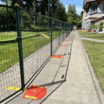 A green construction fence with yellow caution bases and warning signs, positioned along a concrete sidewalk next to a building and a grassy area.