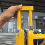 A close-up of a person holding a yellow fence connector, demonstrating how it fits into a metal fence panel, with stacks of fencing materials visible in the background.