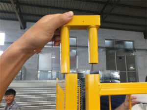 A close-up of a person holding a yellow fence connector, demonstrating how it fits into a metal fence panel, with stacks of fencing materials visible in the background.