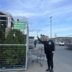 A person in a black sweatshirt standing next to a green fence with a sign that reads "We Rent Fence" from "Sunbelt Rentals," gesturing towards the sign while shopping carts are visible in the background.