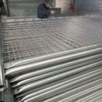 A large stack of newly manufactured, galvanized temporary fence panels with mesh infills, likely in a factory or warehouse. A worker wearing a face mask is visible in the background, working on a panel.