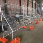 Heavy-duty temporary fence panels with welded mesh and orange plastic water-filled fence feet, featuring stability bracing, set up in an indoor facility.