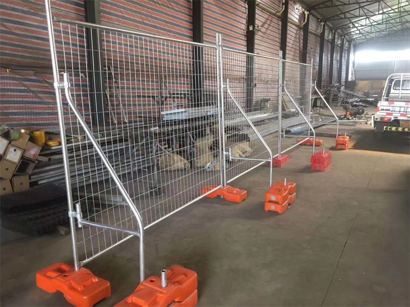 Heavy-duty temporary fence panels with welded mesh and orange plastic water-filled fence feet, featuring stability bracing, set up in an indoor facility.