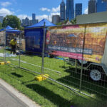 Temporary mesh fencing with yellow plastic bases enclosing a grassy area. In the background, a trailer displays a banner for the "Asia Street Food Festival," with blue tents and a city skyline under a clear sky.