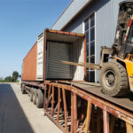 A forklift loading large, flat panels into the back of an open shipping container, which is positioned on a truck backed onto a loading ramp.
