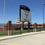 Black steel picket fence with brick pillars securing the perimeter of a large sports stadium or school facility, with a "Yukon Millers" sign and bleachers visible.