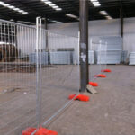 A temporary mesh fence with orange plastic bases set up inside a large industrial warehouse or factory space. A tall metal support pillar stands within the fenced area, and various industrial materials are visible in the background.