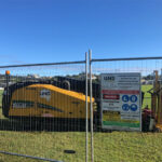 Temporary construction fence enclosing a yellow Vermeer D23X30 S3 drilling machine and an "Underground Network Services" safety sign on a grassy field.