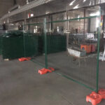 Green powder-coated temporary mesh fencing with orange plastic bases, set up inside a warehouse with stacks of similar green fence panels in the background.