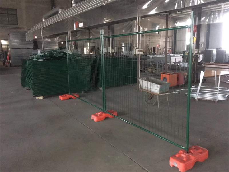 Green powder-coated temporary mesh fencing with orange plastic bases, set up inside a warehouse with stacks of similar green fence panels in the background.