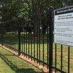 A long black steel picket fence in a green park-like setting, featuring a white sign titled "Maintenance Buden" (likely Maintenance Burden), indicating information about fence upkeep.