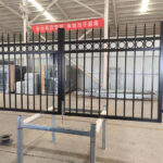 Two individuals are holding open a large, decorative black metal double gate inside a warehouse or factory. The gate features vertical pickets with pointed tops and a horizontal top rail adorned with a series of circular designs. A sign with Chinese characters is visible in the background.