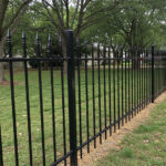 A long black metal fence with pointed finials on its pickets extends through a residential area with a green lawn. Mature trees and houses are visible in the background.
