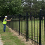 A worker in a hard hat and safety vest is examining a section of a black metal fence that runs alongside a paved sidewalk and a grassy park area.