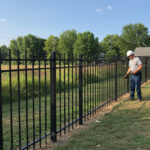 A worker in a white hard hat and blue jeans is inspecting or working on a black metal fence that runs along a grassy area, with a field and trees in the background.