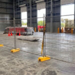 Temporary mesh fencing with yellow plastic bases and support poles set up on a concrete floor inside a large industrial warehouse, with equipment and roll-up doors in the background.