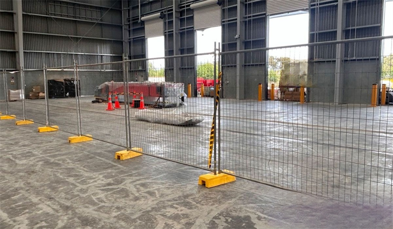 Temporary mesh fencing with yellow plastic bases and support poles set up on a concrete floor inside a large industrial warehouse, with equipment and roll-up doors in the background.