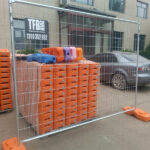 Temporary mesh fencing with orange plastic bases in the foreground, with large stacks of orange plastic bases and a gray car parked behind it, all in front of a building with a "TFB Temporary Fence & Barrier" sign.
