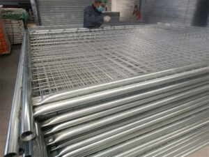 A large stack of newly manufactured, galvanized temporary fence panels with mesh infills, likely in a factory or warehouse. A worker wearing a face mask is visible in the background, working on a panel.