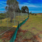 A long line of temporary mesh fencing with yellow bases, and a green silt fence attached along its bottom, stretching across a grassy, undeveloped field.